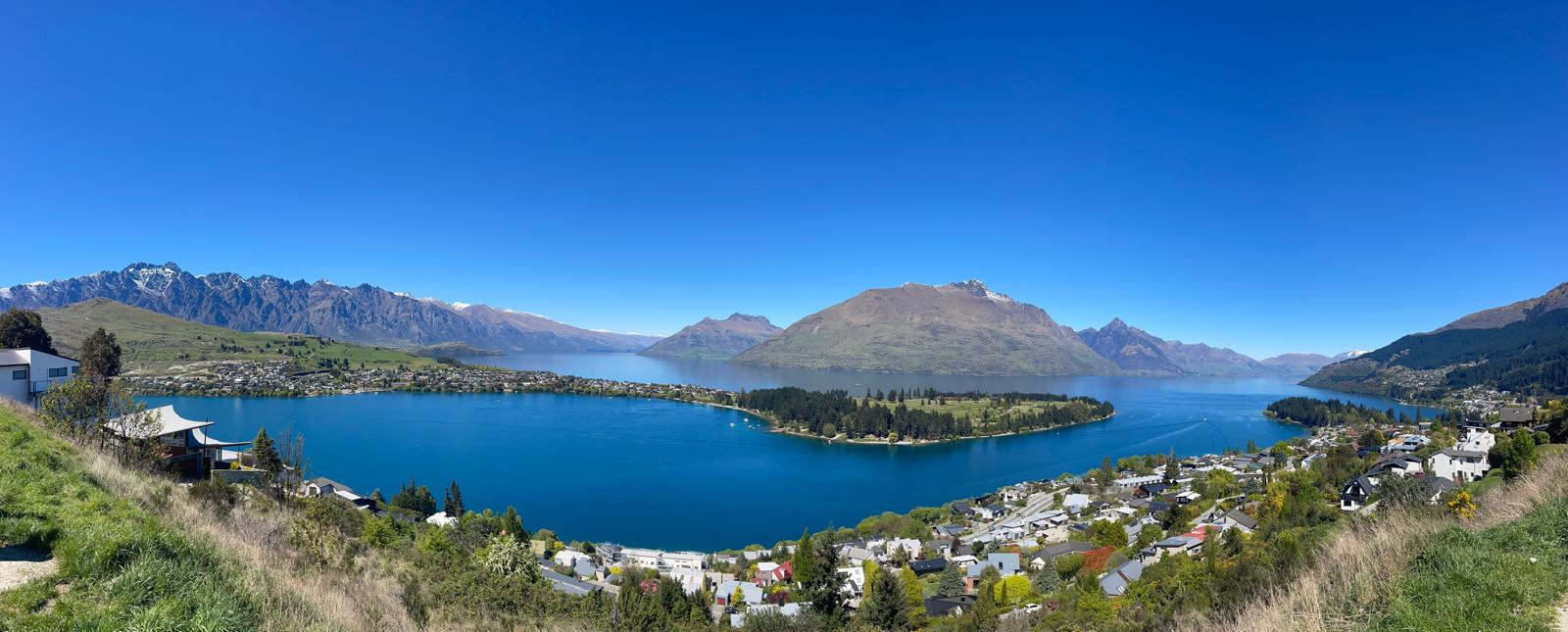 An elevated panorama across a blue lake, mountain ranges and a lakeside settlement.