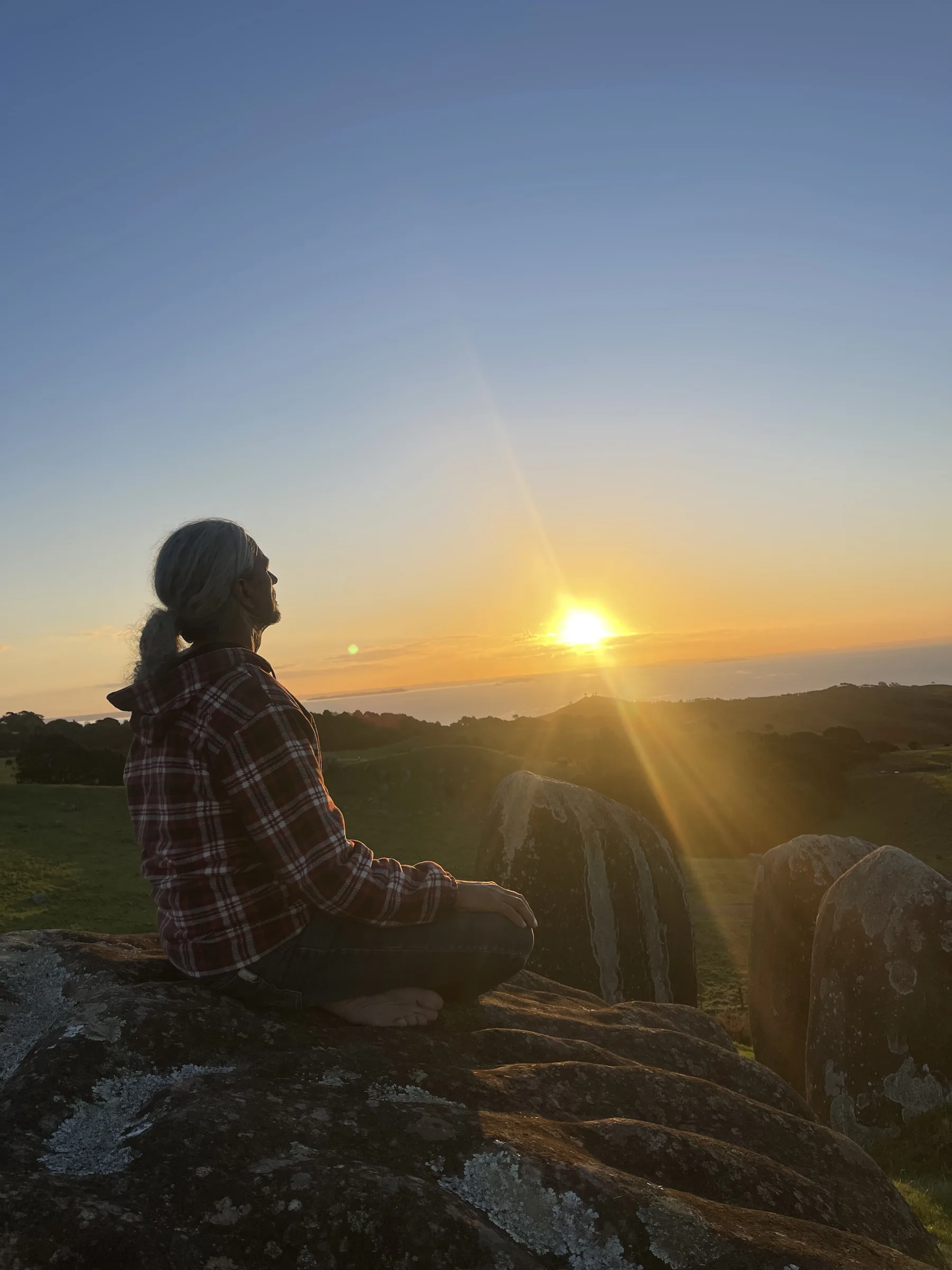 Una persona siede in silenzio su una roccia a Stony Batter, contemplando le colline di Waiheke e il mare nella luce della sera.